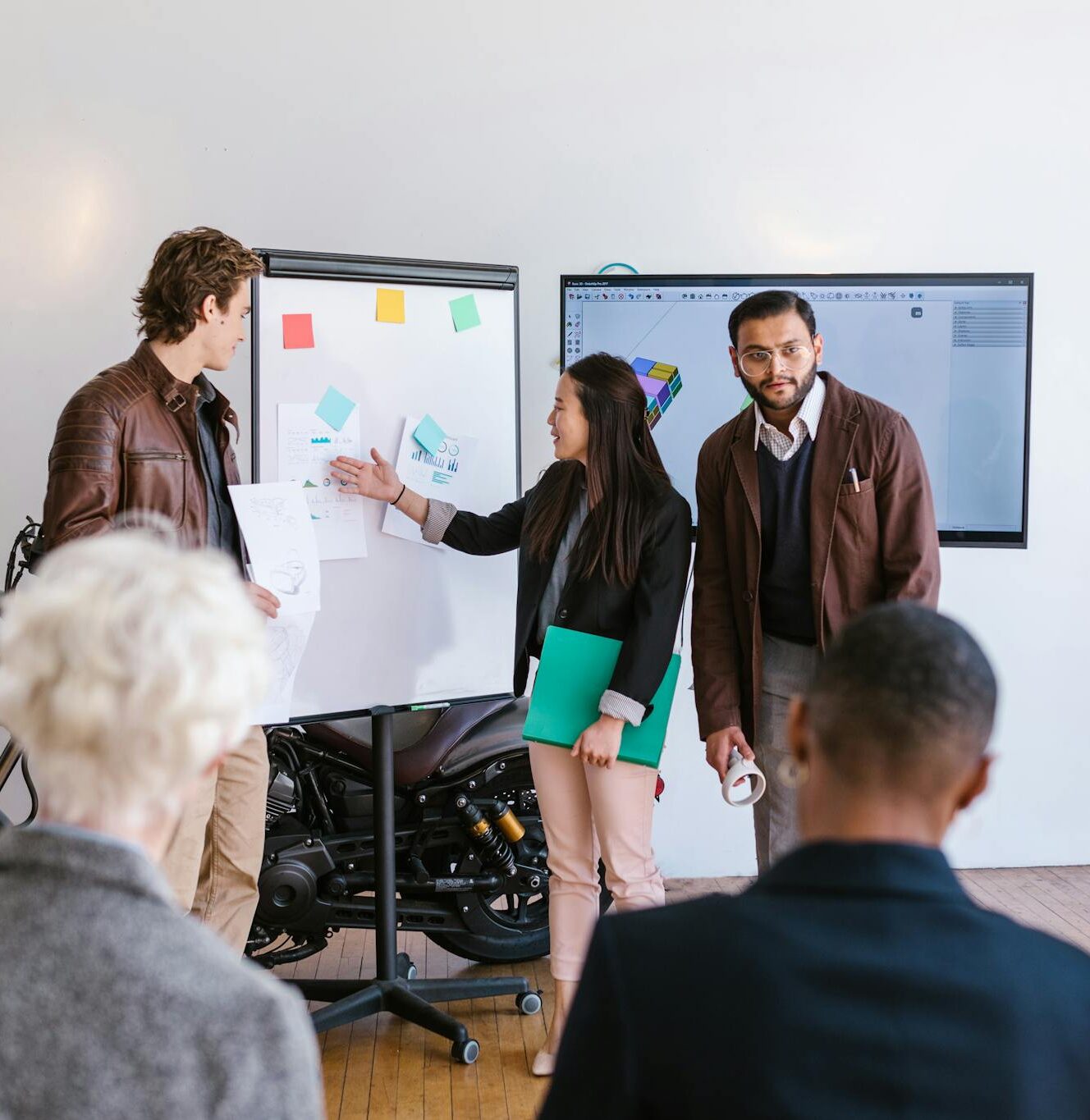 A multicultural team collaborates in an office, presenting ideas using a whiteboard and digital screen.